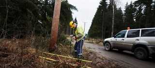 Man i arbetskläder och gul hjälm står vid en elstolpe i skogsmiljö